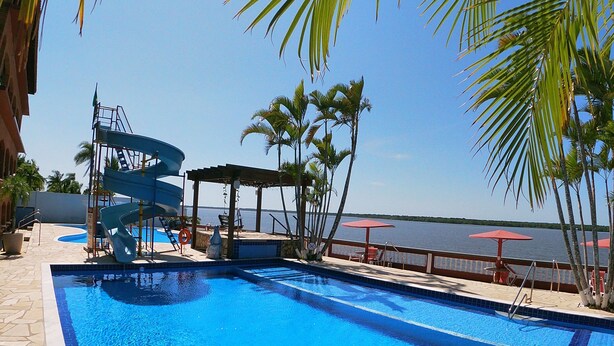 A swimming pool with a water slide, palm trees, and a view of the water. - Hotel Marazul (Cananeia)