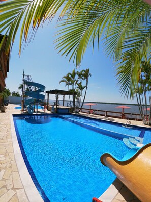 A swimming pool with a water slide, palm trees, and a view of the water. - Hotel Marazul (Cananeia)
