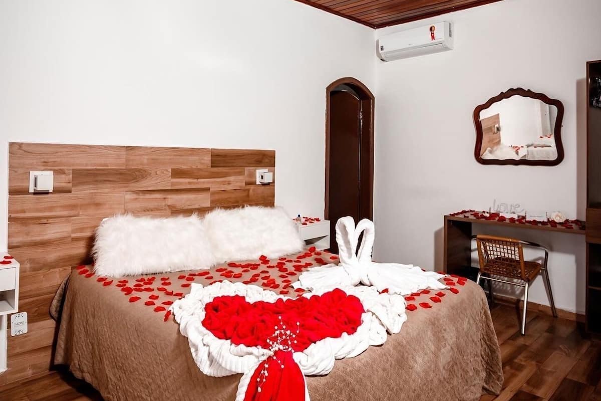 A bedroom with a wooden headboard, a bed with rose petals and a heart-shaped pillow, a mirror, and a desk with a chair.