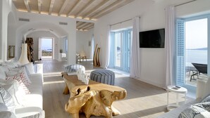 Een ruime woonkamer met een houten salontafel, een bank en een aan de muur bevestigde televisie. Er is een groot raam met luiken en uitzicht op zee.