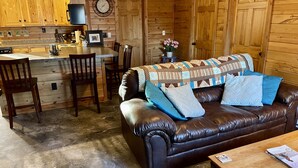 Un accogliente interno di una baita con un divano in pelle, un tavolo da pranzo in legno e un angolo cottura con mobili in legno e una sveglia.