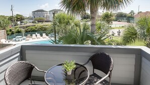 A balcony with two wicker chairs and a small table, a potted plant, and a view of a swimming pool and buildings in the background. - Cozy Condo, 3 Minute Walking Distance to the Beach (Destin)