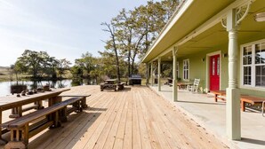 A wooden deck with picnic tables and a barbecue grill, overlooking a lake with trees and a building with a red door.