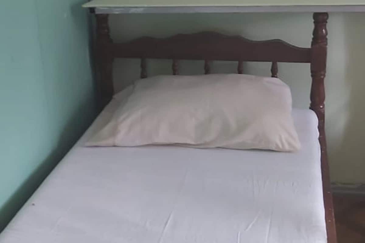 A small, single-bed room with a wooden headboard and a white mattress. There is a shelf above the bed with a waffle-like object and a brown item hanging on it.