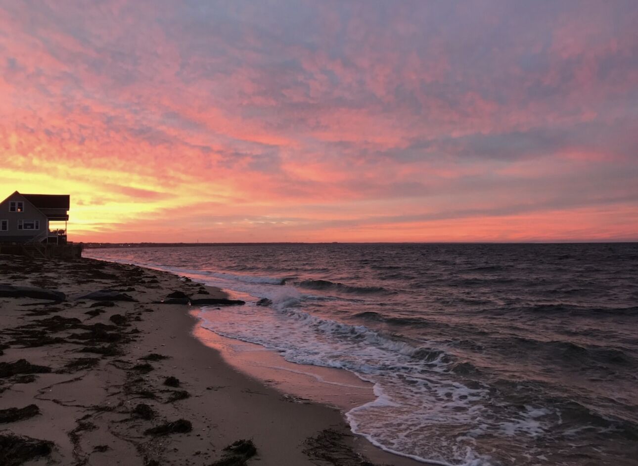 Ein Strand mit Wellen bei Sonnenuntergang, ein Haus links und ein klarer Himmel.