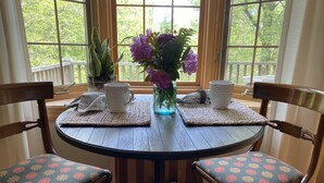 Ein runder Holztisch mit zwei Stühlen, beide mit floraler Polsterung. Auf den Untersetzern stehen zwei weiße Keramiktassen, eine Vase mit lila Blumen und ein Topfpflanze.