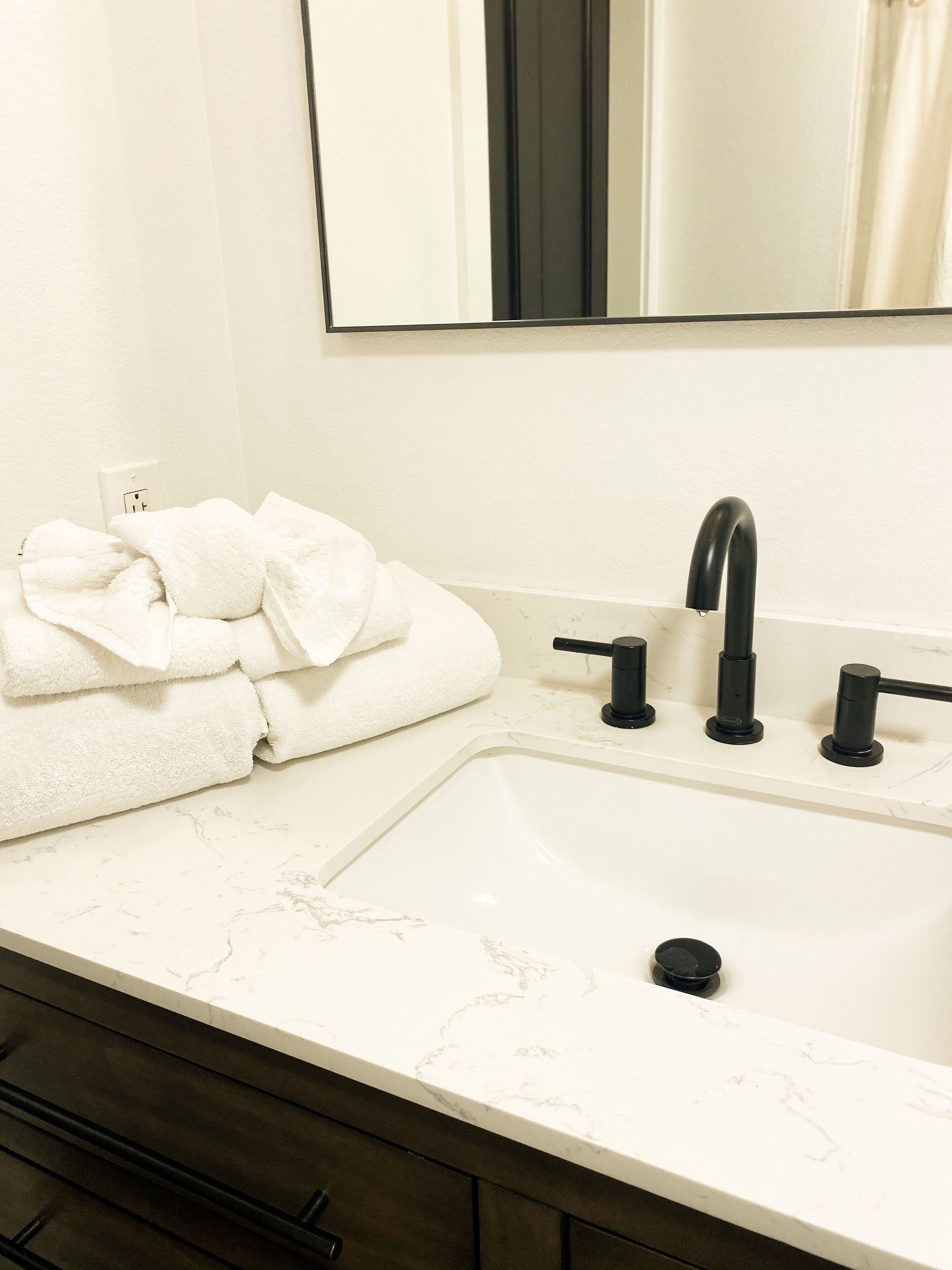 Une salle de bain avec un lavabo blanc, un robinet noir et un miroir au-dessus. Des serviettes sont pliées et empilées sur le côté gauche du lavabo.