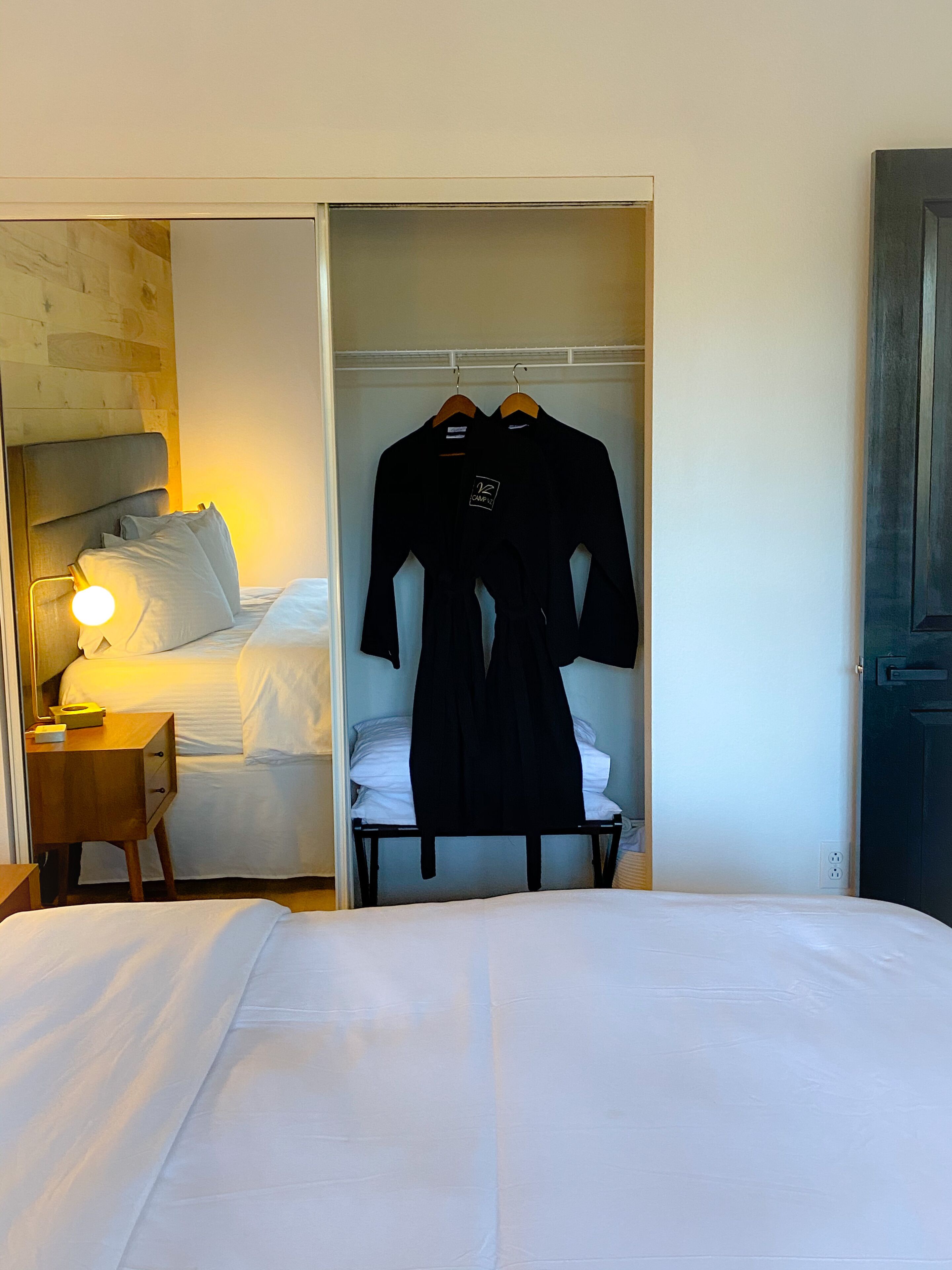 A hotel room with a bed, a bedside table with a lamp, a wardrobe, and two black robes hanging on a rack.