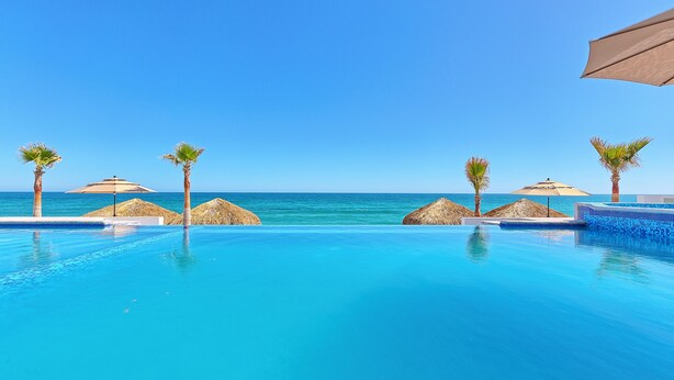 A clear blue pool with a view of the ocean, surrounded by palm trees and thatched umbrellas. - Luxury Beachfront Six Palms Villas # 2 (puerto penasco)