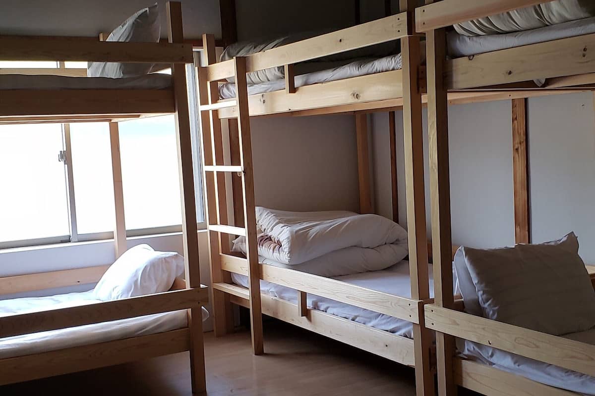 A room with three bunk beds, wooden flooring, a hanging light fixture, and a window providing natural light.