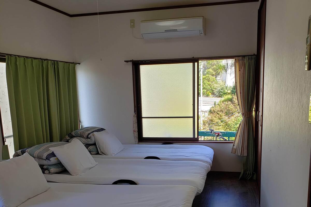 A hotel room with three single beds, a window with a view of greenery, an air conditioning unit, and a small desk with a computer.