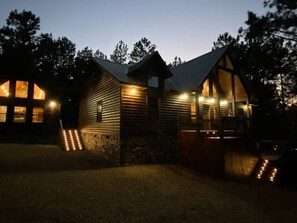 Una cabaña de troncos con cimientos de piedra, techo de metal y un porche iluminado.
