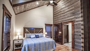 Un dormitorio con techo de madera, una cama con ropa de cama a rayas, dos mesitas de noche con lámparas y una puerta que da a otra habitación.