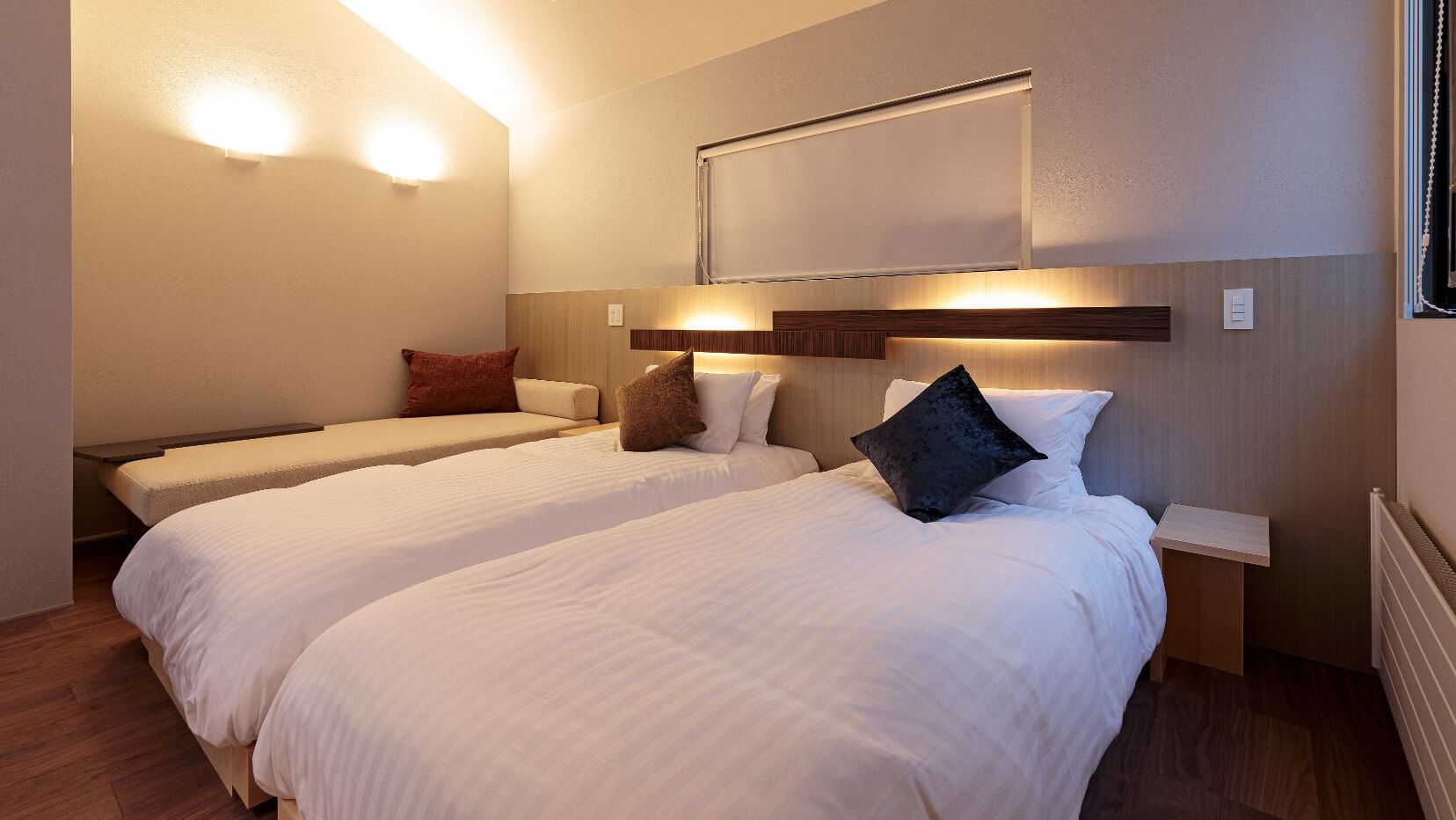 A hotel room with two beds, a nightstand, a wall-mounted light, and a window with a curtain.