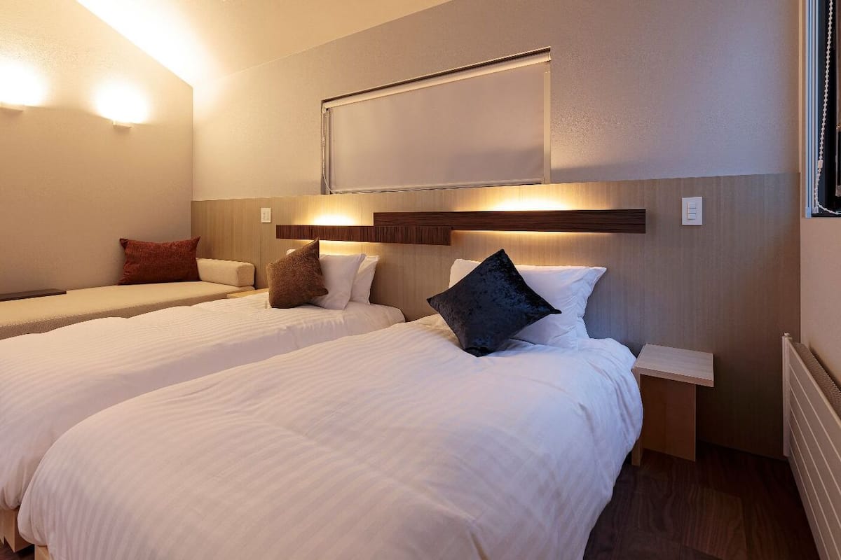A hotel room with two beds, a nightstand, a wall-mounted light, and a window with a curtain.