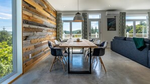 Ein stilvolles Esszimmer mit Holztisch und Stühlen, großen Fenstern mit Blick ins Grüne und einem grauen Sofa im Hintergrund.