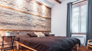 Una camera da letto con parete in mattoni, struttura letto in legno, un tavolino con una pianta e una finestra con persiane.