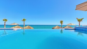 Una piscina azul cristalina con vistas al océano, rodeada de palmeras y sombrillas de paja.