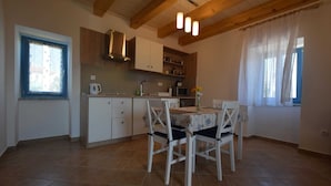 A kitchen with white cabinets, a dining table with chairs, a window with curtains, and a hanging light fixture. - Studio apartment Depope 2 (Skrbčići)
