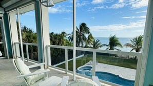 Un'area esterna riparata con arredi bianchi, una piscina e vista sull'oceano e sulle palme.