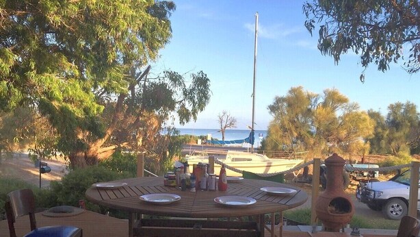 An outdoor dining area with a round wooden table and chairs, surrounded by trees and a view of the sea.