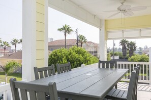 A covered outdoor patio with a table and chairs, a ceiling fan, and a view of surrounding houses and greenery.