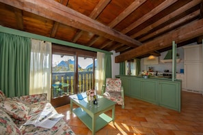 A room with a wooden ceiling, a sofa, a table with a book and glasses, a chair, a kitchen area, and a view of mountains through a window.