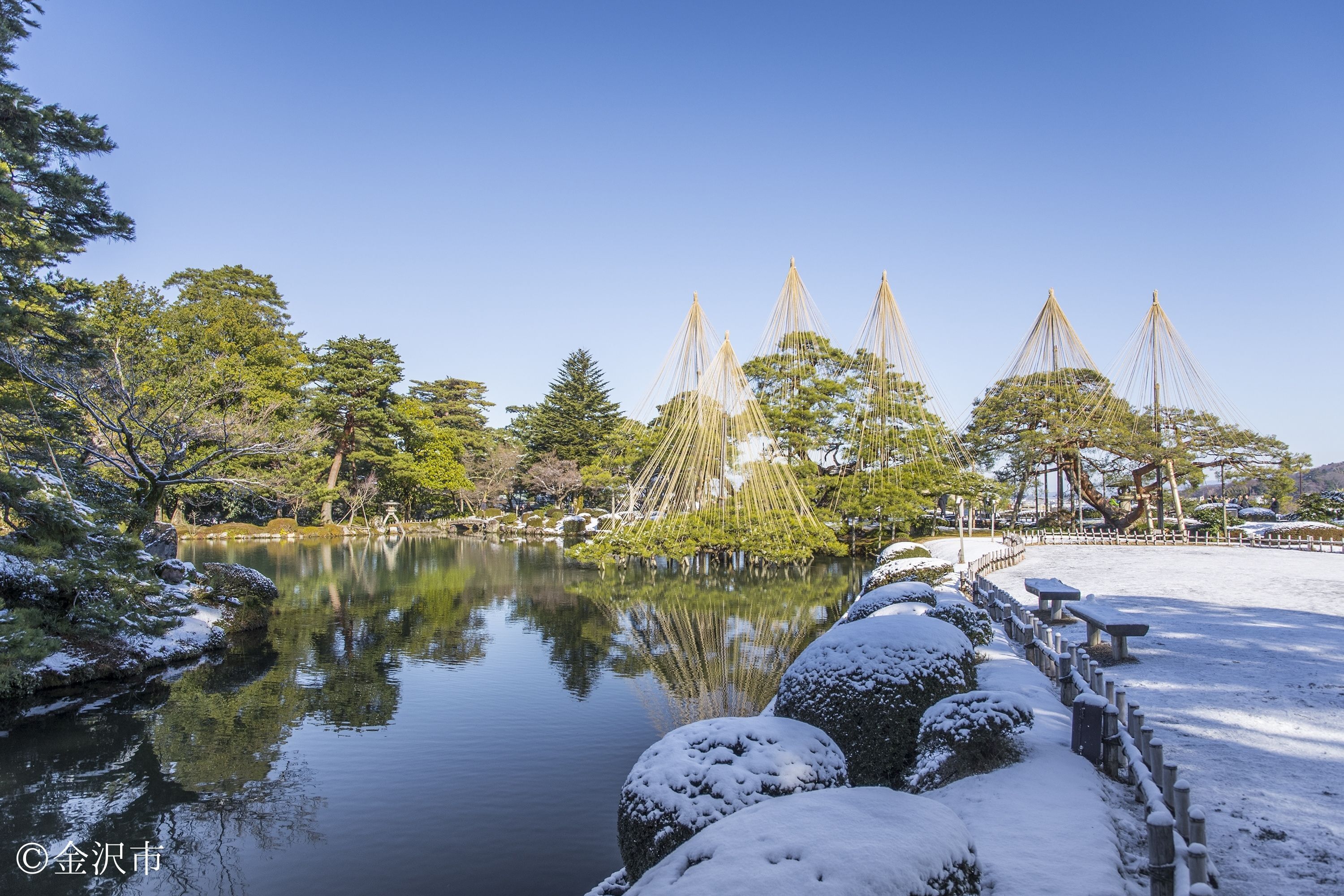 A serene park with a snow-covered pathway, a calm lake, and traditional Japanese structures.