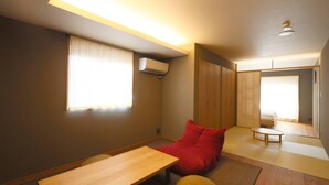 A compact room with a futon bed, a small wooden table, and a futon chair. There is a window with curtains, an air conditioning unit mounted on the wall, and a ceiling light fixture. - NEON HOTEL (Kanazawa)