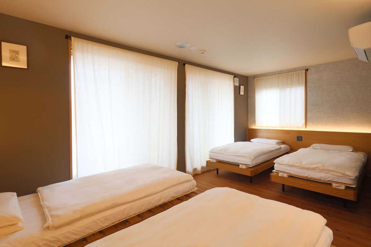 A hotel room with three single beds, a wooden floor, and large windows with white curtains.