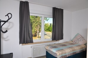 A bedroom with a bed, a radiator, a window with curtains, and a view of a road and trees outside. - Weisenau Plaza (Mainz)