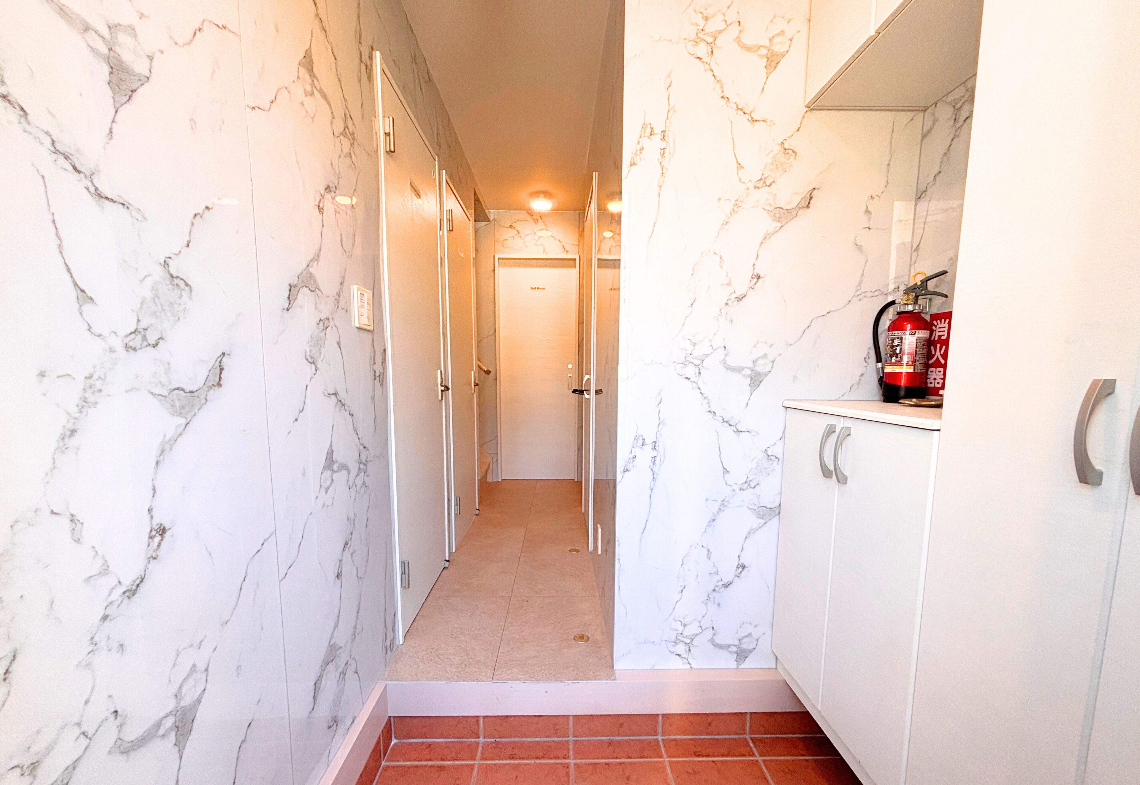 A hallway with marble walls and a tiled floor leading to a door. A fire extinguisher is mounted on the wall.