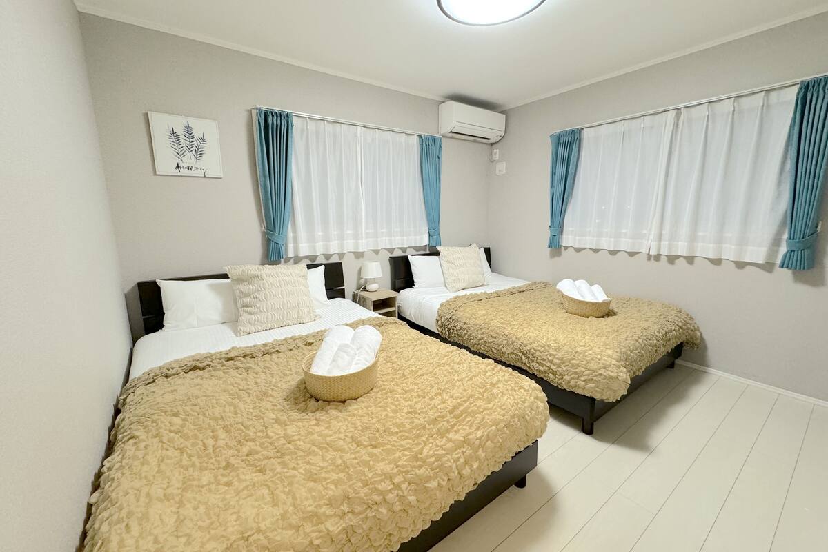 Une chambre d’hôtel avec deux lits, chacun recouvert d’une courtepointe beige à texture et agrémenté d ’un panier rempli de serviettes pliées. Un climatiseur mural est fixé au mur, et une œuvre d’art encadrée est accrochée au-dessus des lits.