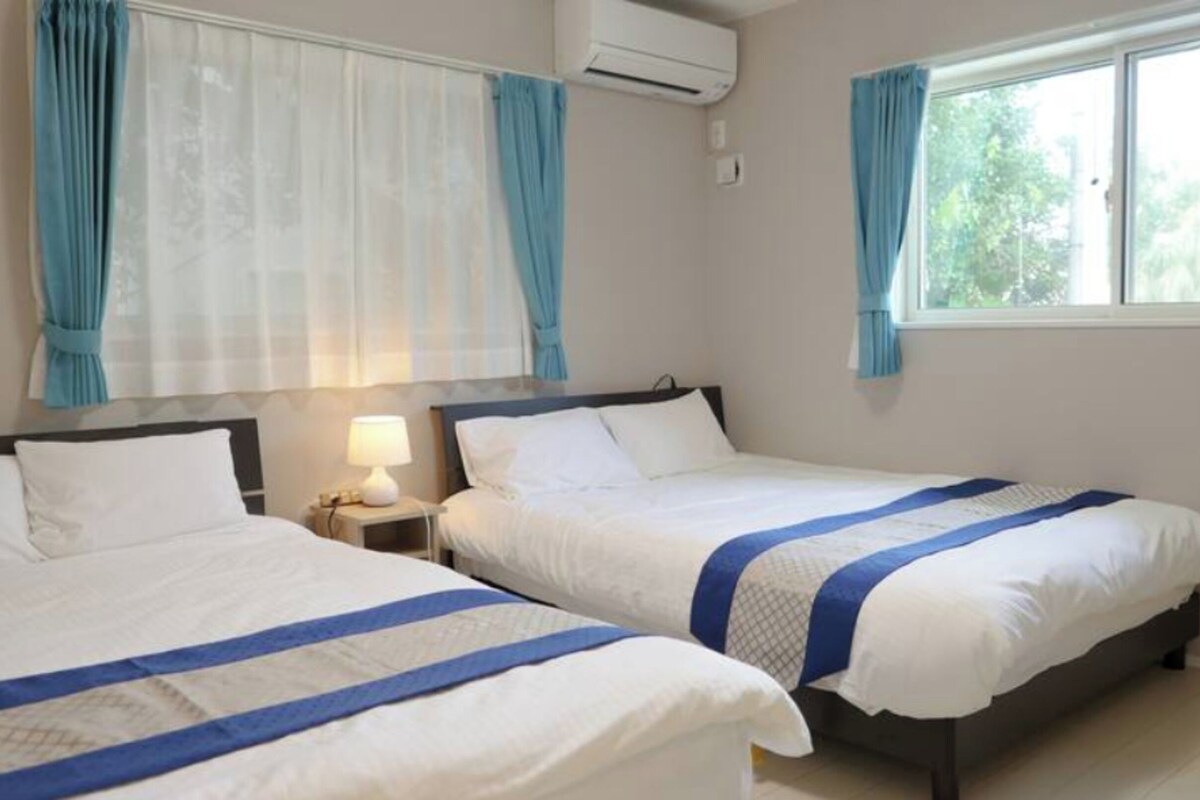 A hotel room with two beds, a nightstand with a lamp, an air conditioning unit, and a window with curtains.