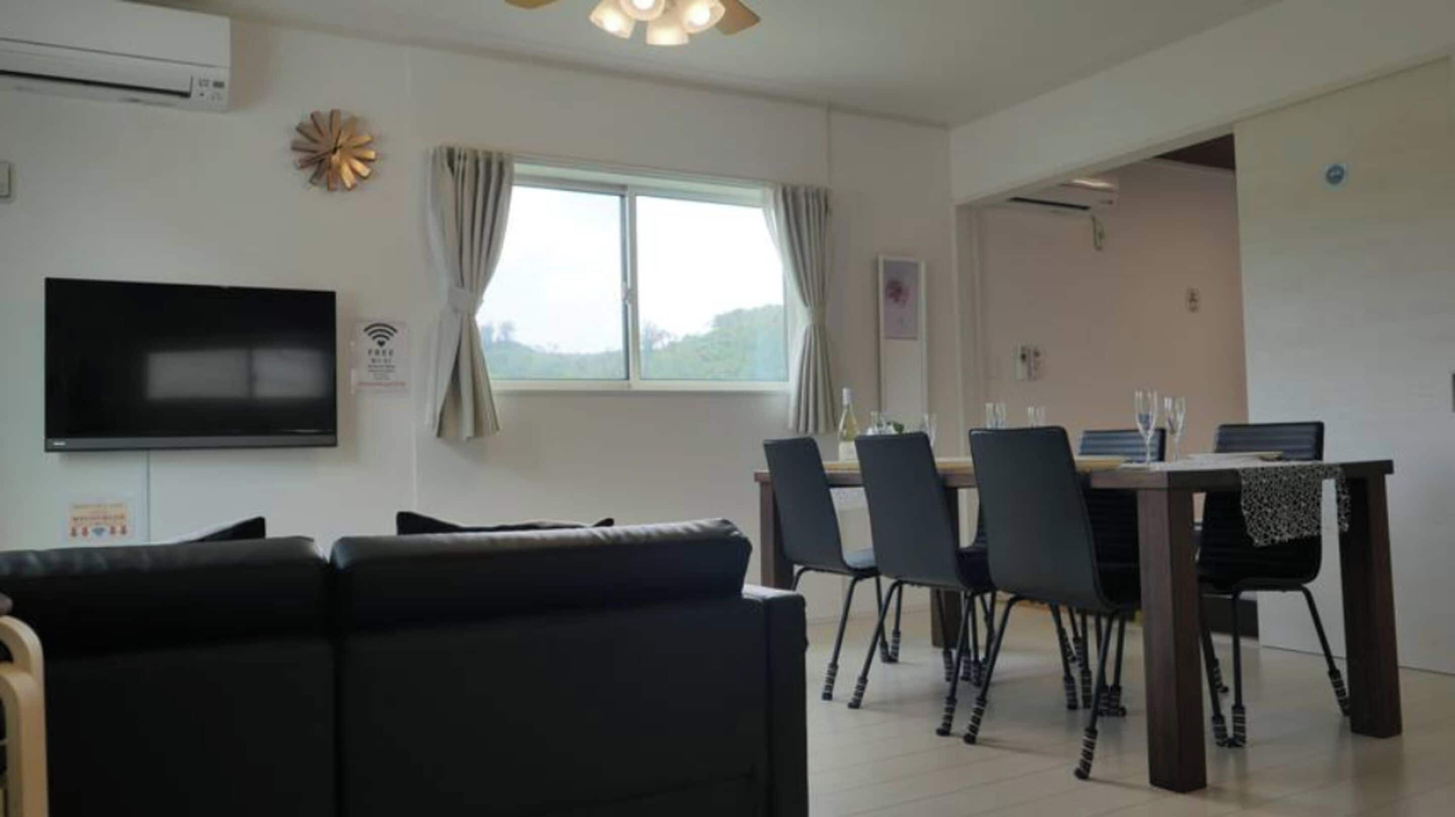 Una sala moderna con un sofá negro, una mesa de comedor con sillas, un televisor de pantalla plana y un equipo de aire acondicionado.