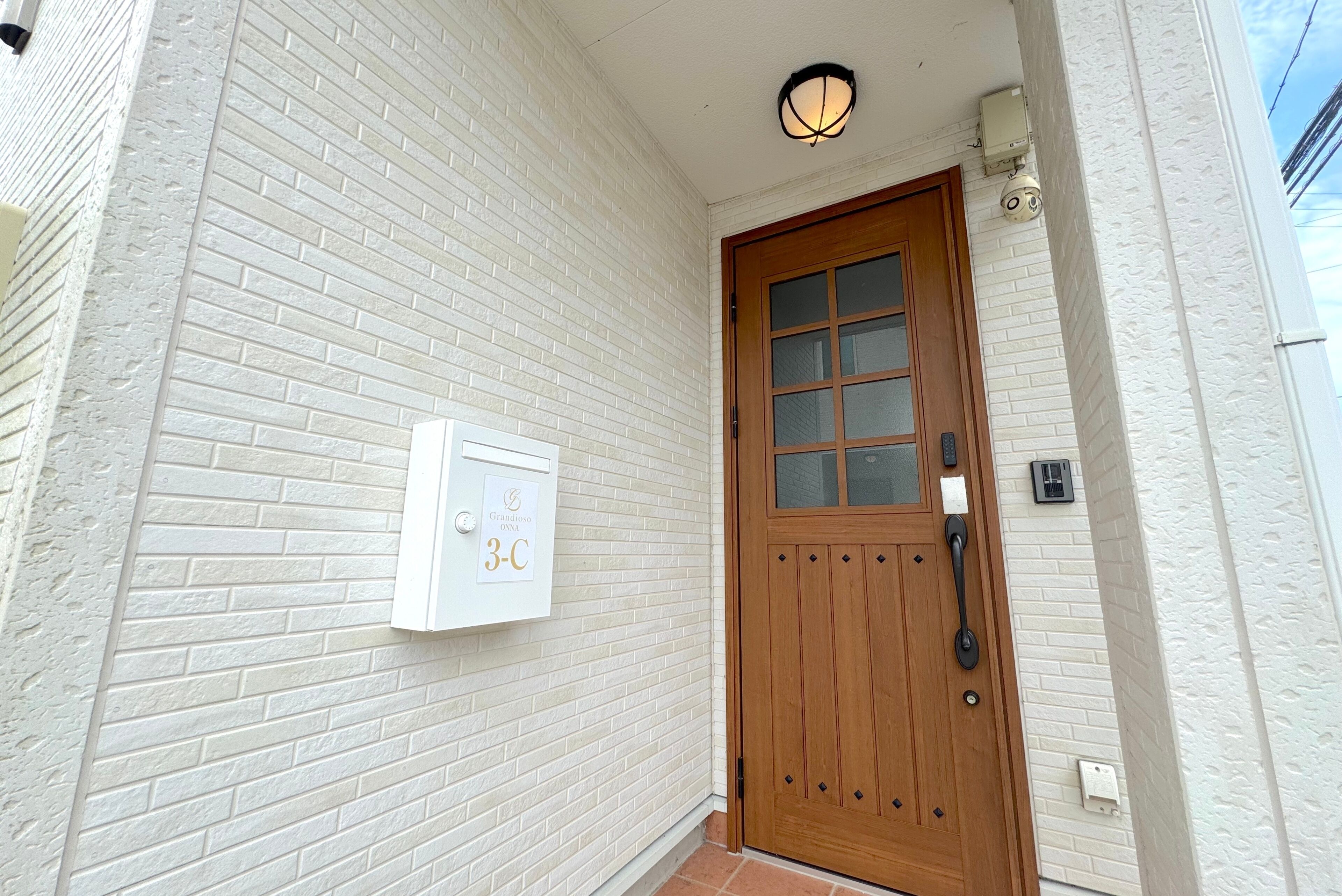 A white brick exterior with a wooden door, a mailbox, and a wall-mounted light fixture.