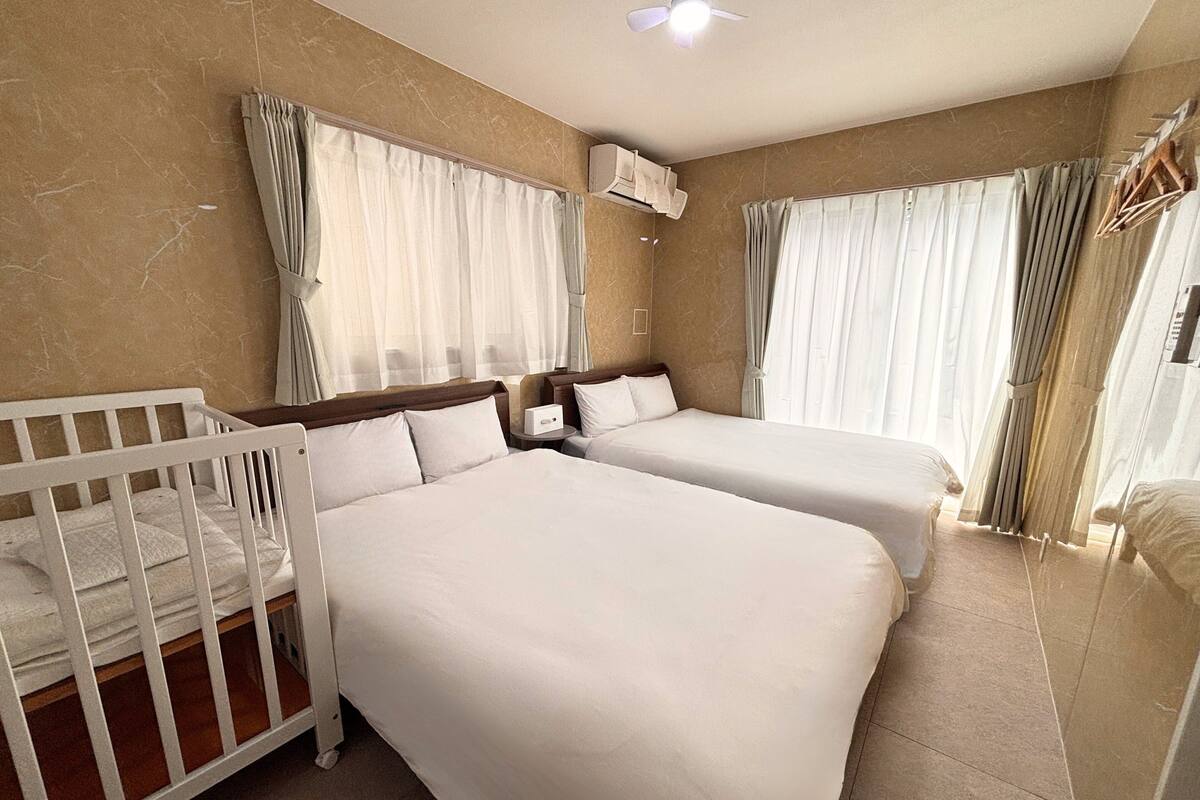 Une chambre d’hôtel avec deux lits, un berceau, un ventilateur de plafond et une fenêtre avec des rideaux.