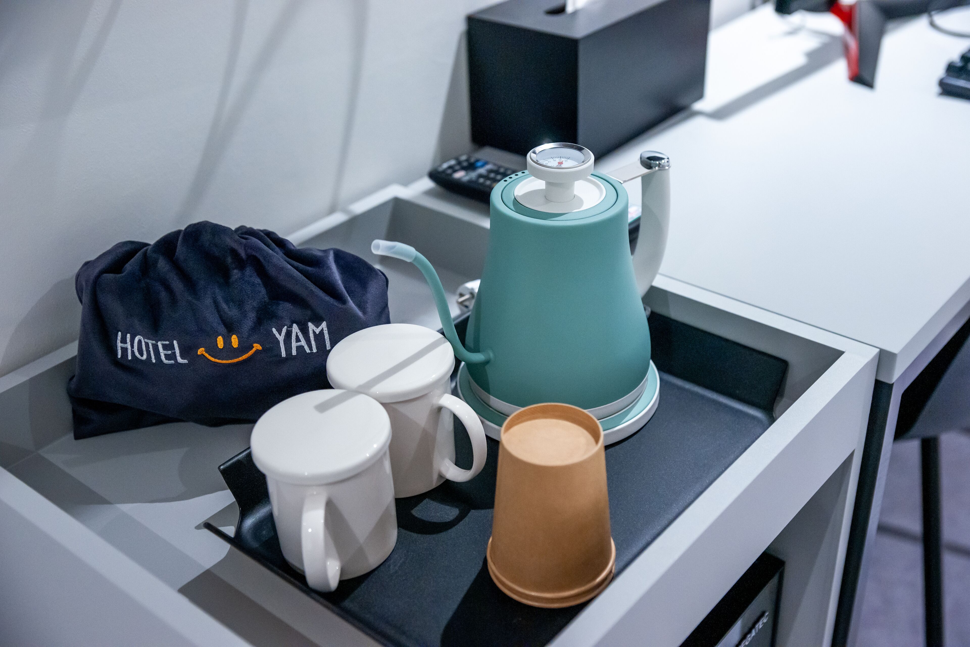 Une chambre d’hôtel équipée d’une machine à café, de deux tasses blanches, d’un gobelet en papier marron et d’un sac bleu sur lequel est inscrit le nom « HOTEL YAM ».