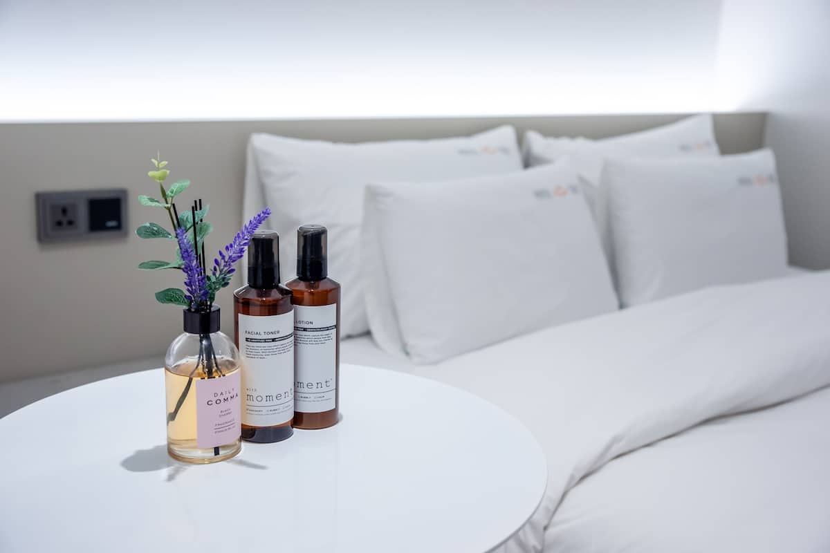Un conjunto ordenado de productos de cuidado de la piel sobre una mesita redonda en una habitación de hotel, con una cama y almohadas blancas al fondo.