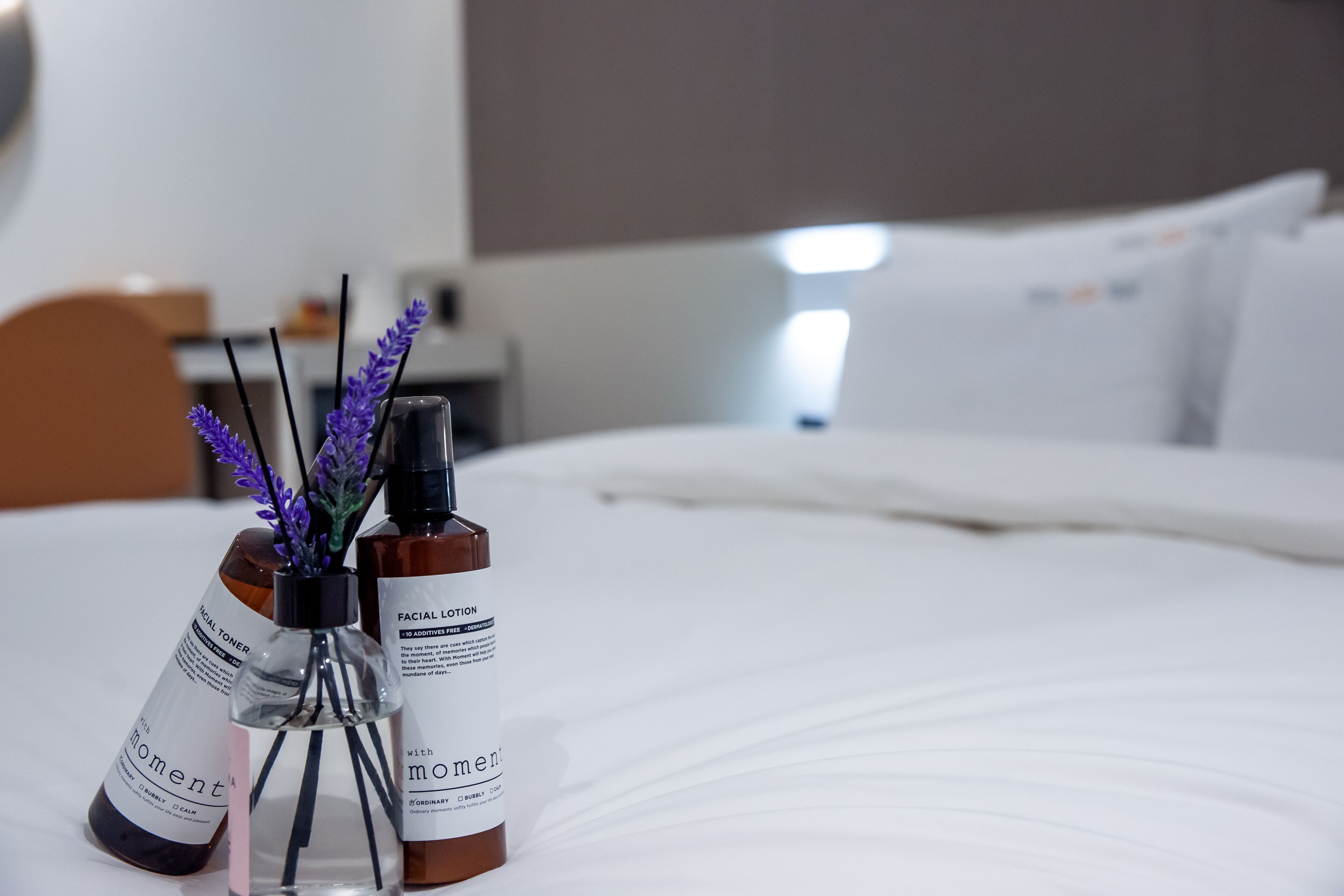 A neatly made bed with a white comforter and pillows, a small table with a vase of lavender, and two bottles of facial lotion.