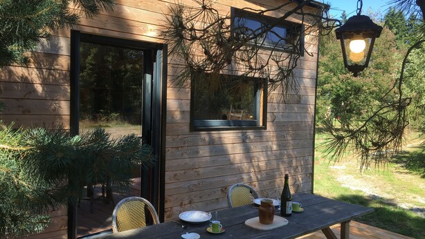 A wooden cabin with a deck, a table with outdoor seating, and a bottle of wine. - La pause Tiny: green relaxation in Morbihan (Ploërmel - (56800))