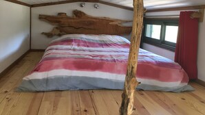 A bedroom with a sloped ceiling, wooden beams, a large bed with a striped blanket, and a window with red curtains. - La pause Tiny: green relaxation in Morbihan (Ploërmel - (56800))