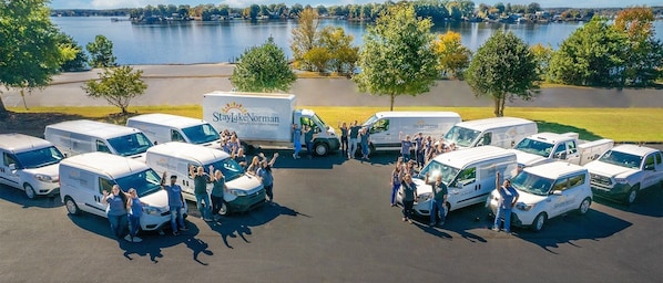 Eine Gruppe von Menschen steht vor mehreren Vans von StayLakeNorman Luxury Vacation Homes, die auf einem Parkplatz geparkt sind. Im Hintergrund sind ein See und Bäume zu sehen.