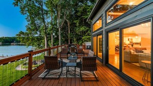 Une terrasse dotée de mobilier d’extérieur, offrant une vue sur un lac, et un bâtiment avec de grandes fenêtres.