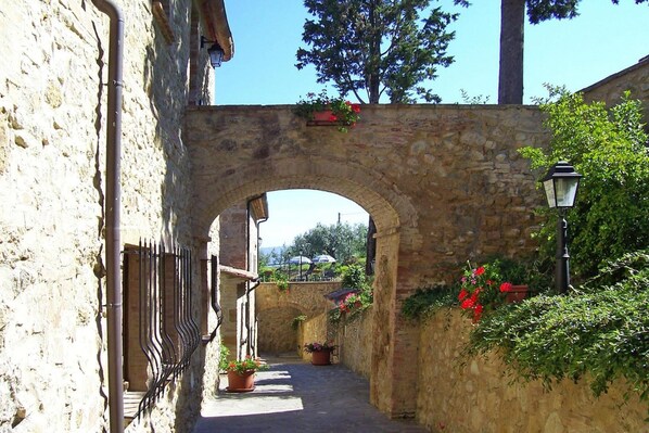 A stone archway leading to a narrow alley with potted plants and a lamp post.