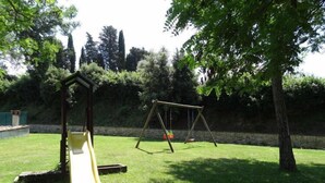 A playground with a slide and swings in a grassy area with trees and a stone wall in the background.
