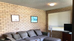 Un salon avec un mur en briques, un canapé gris, une chaise en bois, un téléviseur et une fenêtre avec des stores.