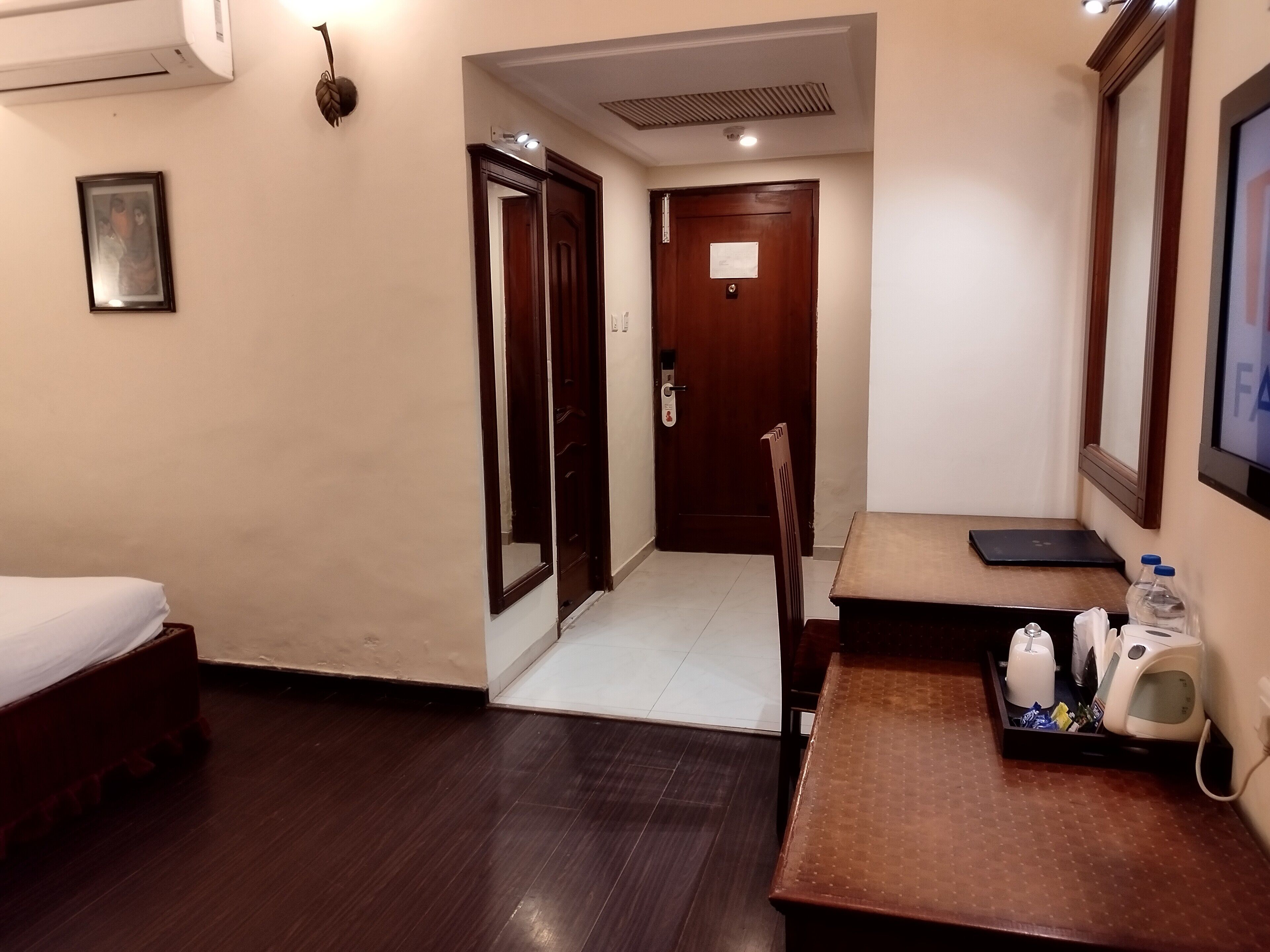 A hotel room with a bed, a desk with a chair, a small table with a tray of toiletries, a wall-mounted TV, and a door leading to another room.