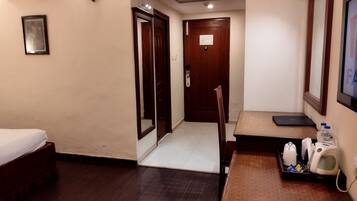 A hotel room with a bed, a desk with a chair, a small table with a tray of toiletries, a wall-mounted TV, and a door leading to another room.