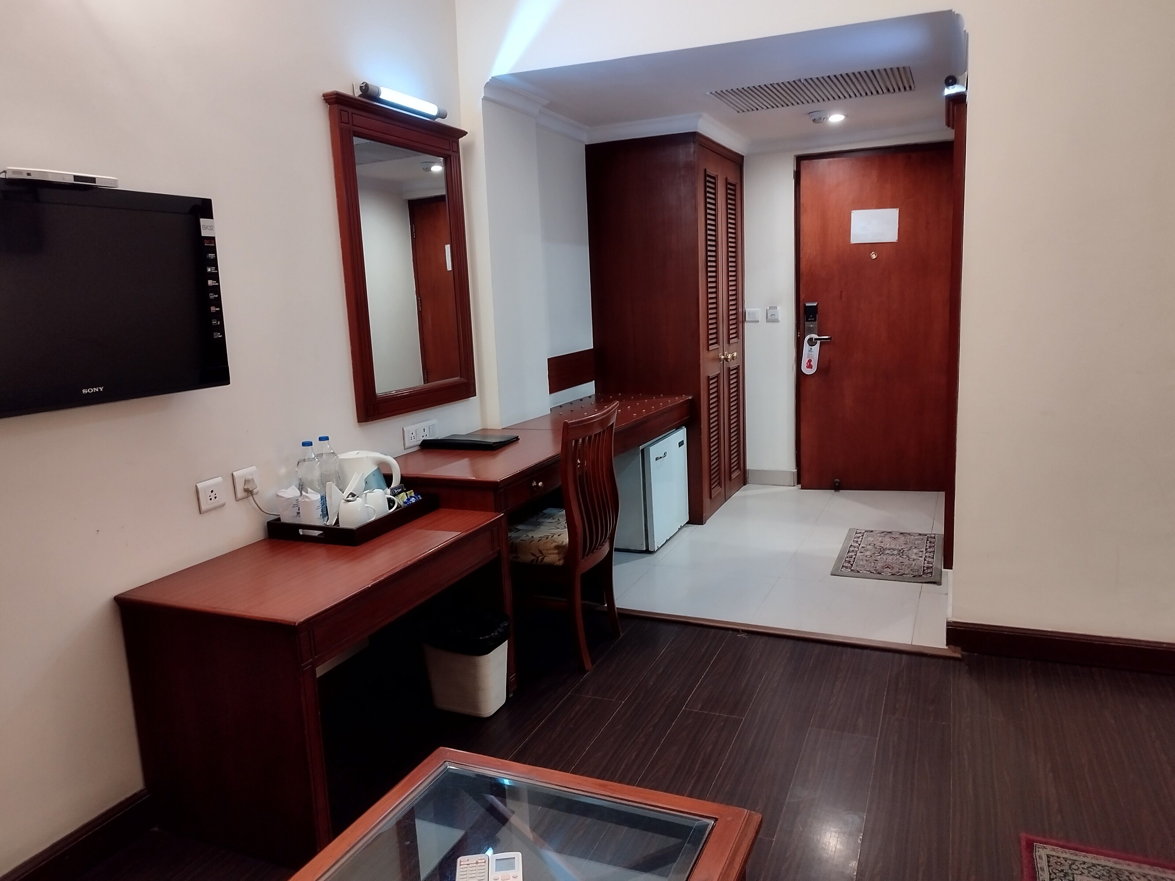 A hotel room with a wooden desk, a flat-screen TV, a mirror, a mini fridge, and a small seating area.